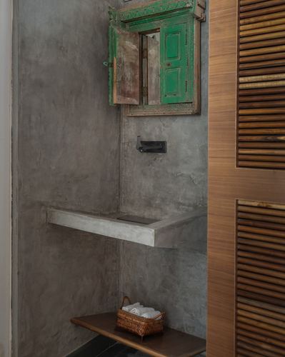 Concrete washroom niche with antique green-shuttered mirror, wall shelf, and wicker towel basket beside a slatted wood door