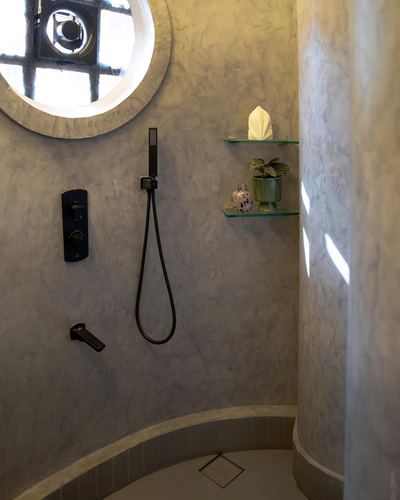 Curved lime-plaster shower stall lit by a round window, with black fixtures and glass shelves.