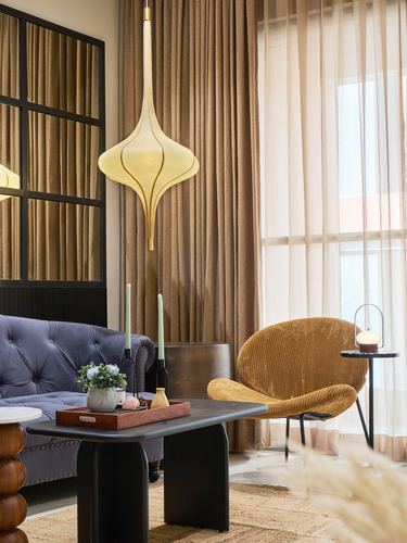 Living room corner with tufted sofa, sculptural pendant light, and curved mustard chair by sheer curtains
