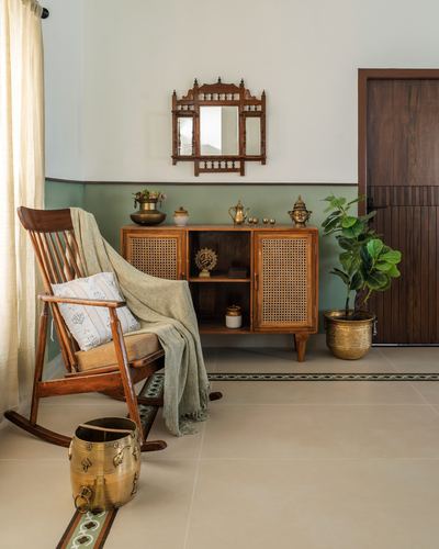Rocking chair with throw beside a cane-front wooden cabinet under a carved mirror, sage dado wall, brass urli and planter, tiled floor with inlaid border.