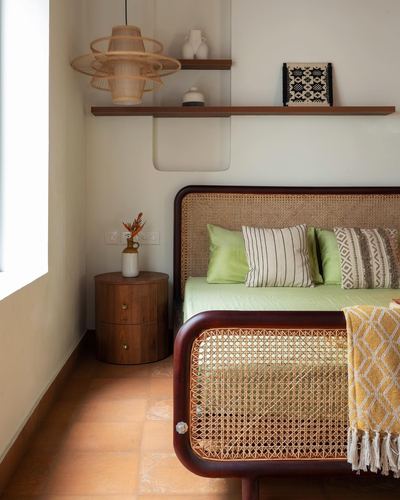 Bedroom corner with curved cane bed, green linens, terracotta floor, and floating wood shelves lit by side window