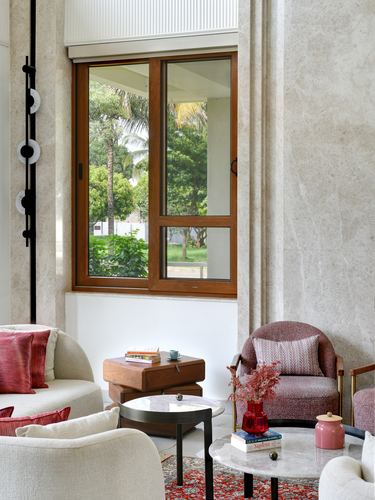 Living room corner with cream sofas, pink armchair, marble tables, and a wooden window overlooking greenery