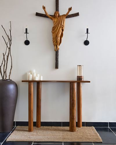Wooden crucifix above a console table with candles, sconces and a vase on black tile floor