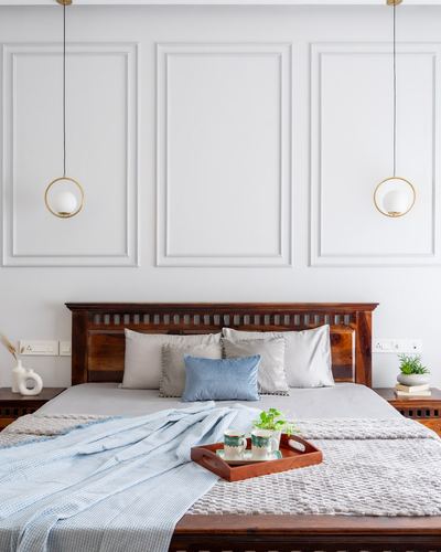 Bedroom with wood headboard, grey and blue linens, wood tray, and two round pendant lights against panelled white wall