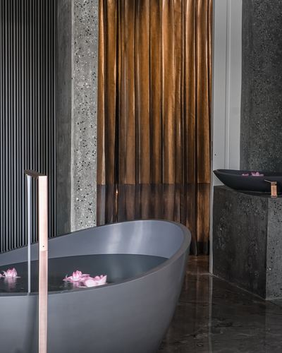 Grey sculptural bathtub with floating petals beside a bronze silk curtain and stone pedestal sink