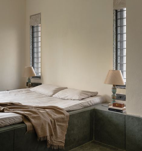 Green stone platform bed with linen bedding beneath narrow jaali windows and turned lamps