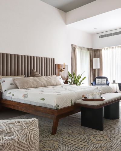 Bedroom with channel-tufted taupe headboard, wooden bed, patterned quilt, bench and floor lamp by curtained window