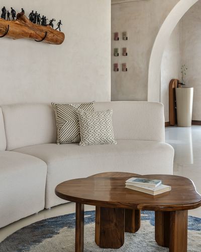 Curved cream sofa with patterned cushions beside a sculptural wooden coffee table in a plastered living room