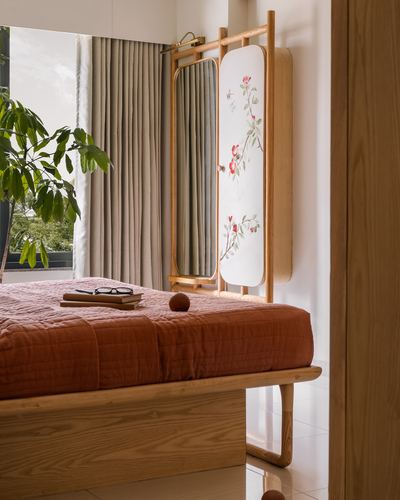 Bed with terracotta quilt beside a wooden-framed mirror and floral folding screen near a curtained window