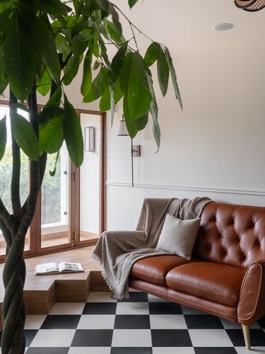 Tufted brown leather sofa beside a checkerboard floor and large indoor plant near a window nook