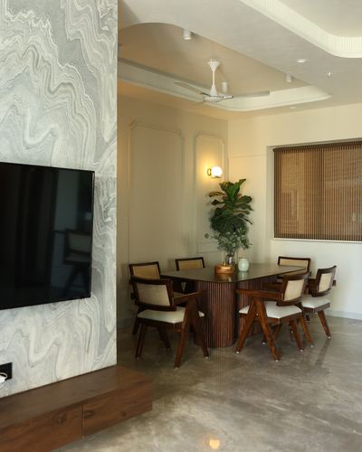 Dining area with fluted-base table, cane chairs, and marble-clad TV wall in Mumbai apartment