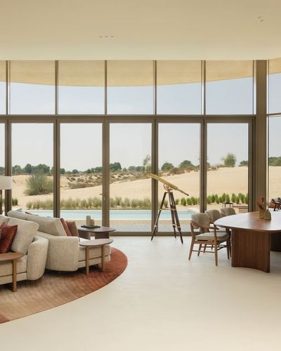 Curved living room with floor-to-ceiling windows overlooking a pool and desert dunes, telescope on tripod