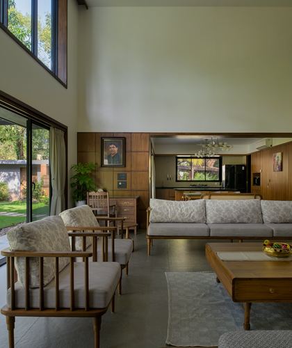 Double-height living room with wood-paneled wall opening to kitchen, cane-backed sofas, sliding glass to garden