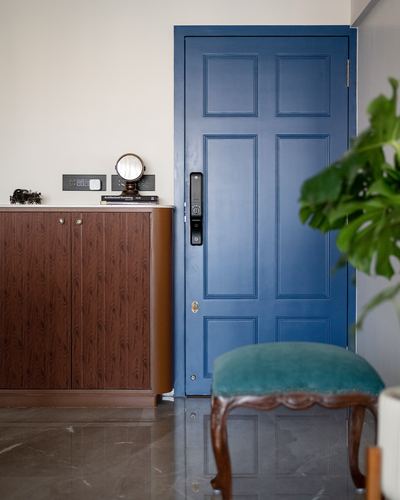 Entry foyer with blue paneled door, wooden cabinet, mirror stand, and teal velvet stool on dark stone floor