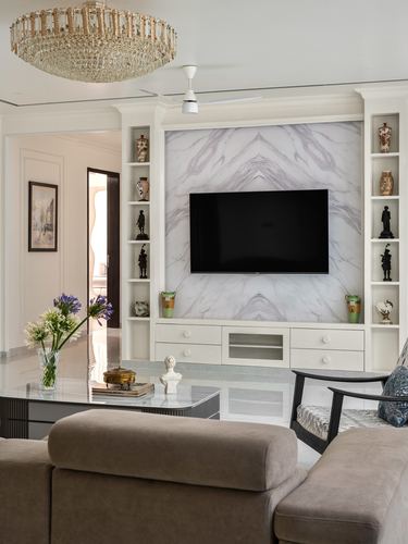 Living room with marble feature wall behind TV, flanked by shelved niches, crystal chandelier, and sofa in foreground