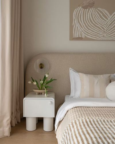 Beige bedroom corner with arched boucle headboard, round nightstand, tulips, and striped artwork