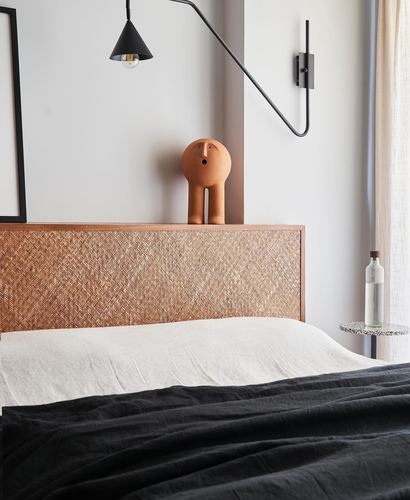 Bedroom corner with woven cane headboard, terracotta figurine, black swing-arm lamp and dark linen bedding