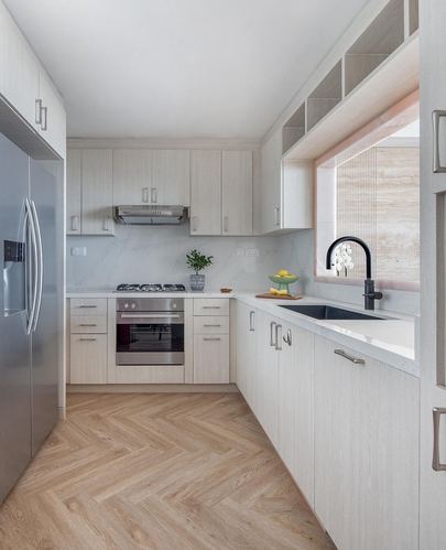 U-shaped minimalist kitchen with light wood cabinetry, white countertop, herringbone floor and stainless appliances