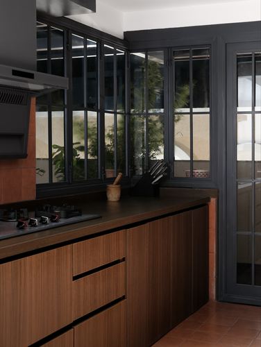 Corner kitchen with wood cabinetry, dark countertop, terracotta floor, and black steel-framed windows overlooking greenery