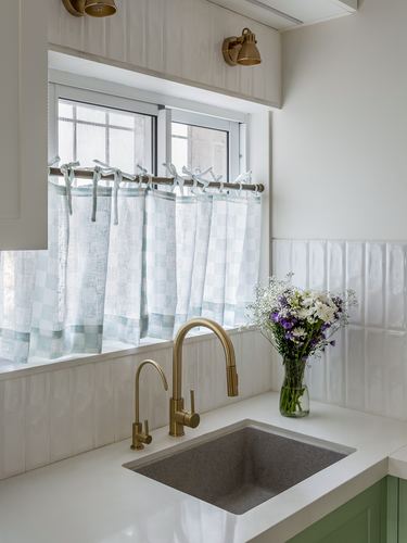 Kitchen sink area with brass faucets, checkered curtains, and a flower vase on white counter
