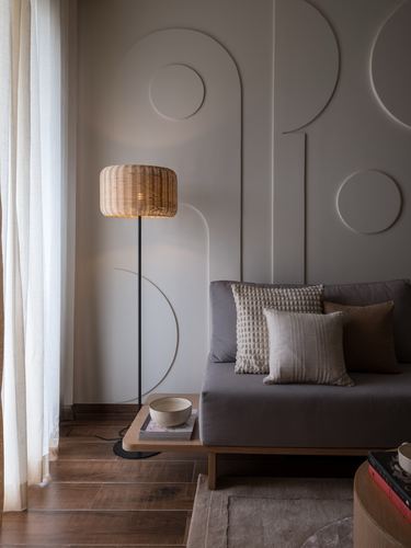 Living room corner with rattan floor lamp, sculpted arch wall relief, and grey sofa with cushions.