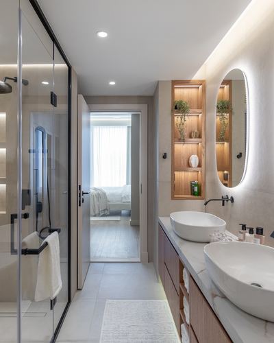 Narrow hotel-style bathroom with glass shower, wood vanity with two vessel sinks, and view through open door to bedroom