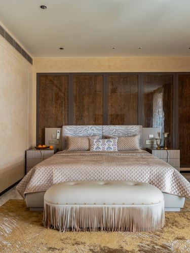 Symmetrical bedroom with quilted bedspread, patterned headboard, and fringed bench against stone-paneled wall