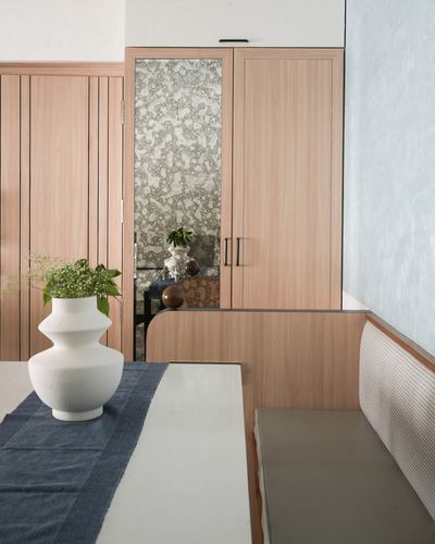 Wooden cabinetry with mirrored insert beside a banquette and a table with a bulbous white vase.