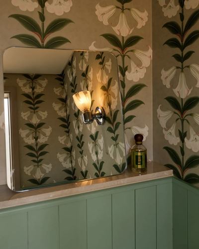 Bathroom corner with floral wallpaper, lit sconce, mirror reflection, marble counter and green cabinetry
