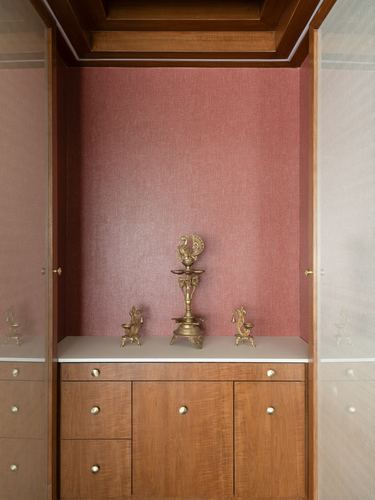 Pooja niche with pink textured wall, brass diyas, and wooden cabinetry below with brass knobs