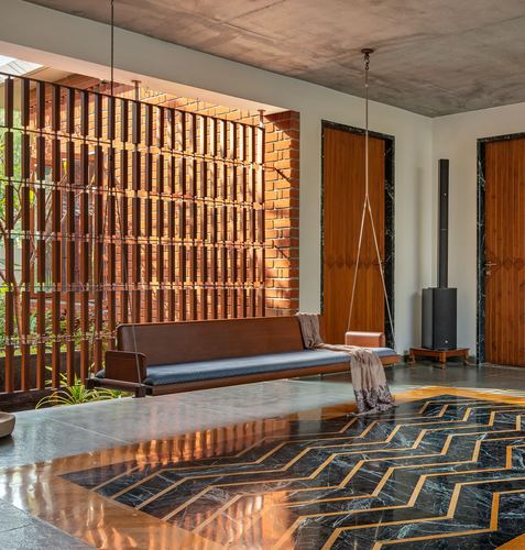 Wooden hanging swing bench before a brick jaali screen, with chevron-patterned marble floor and carved wooden doors.