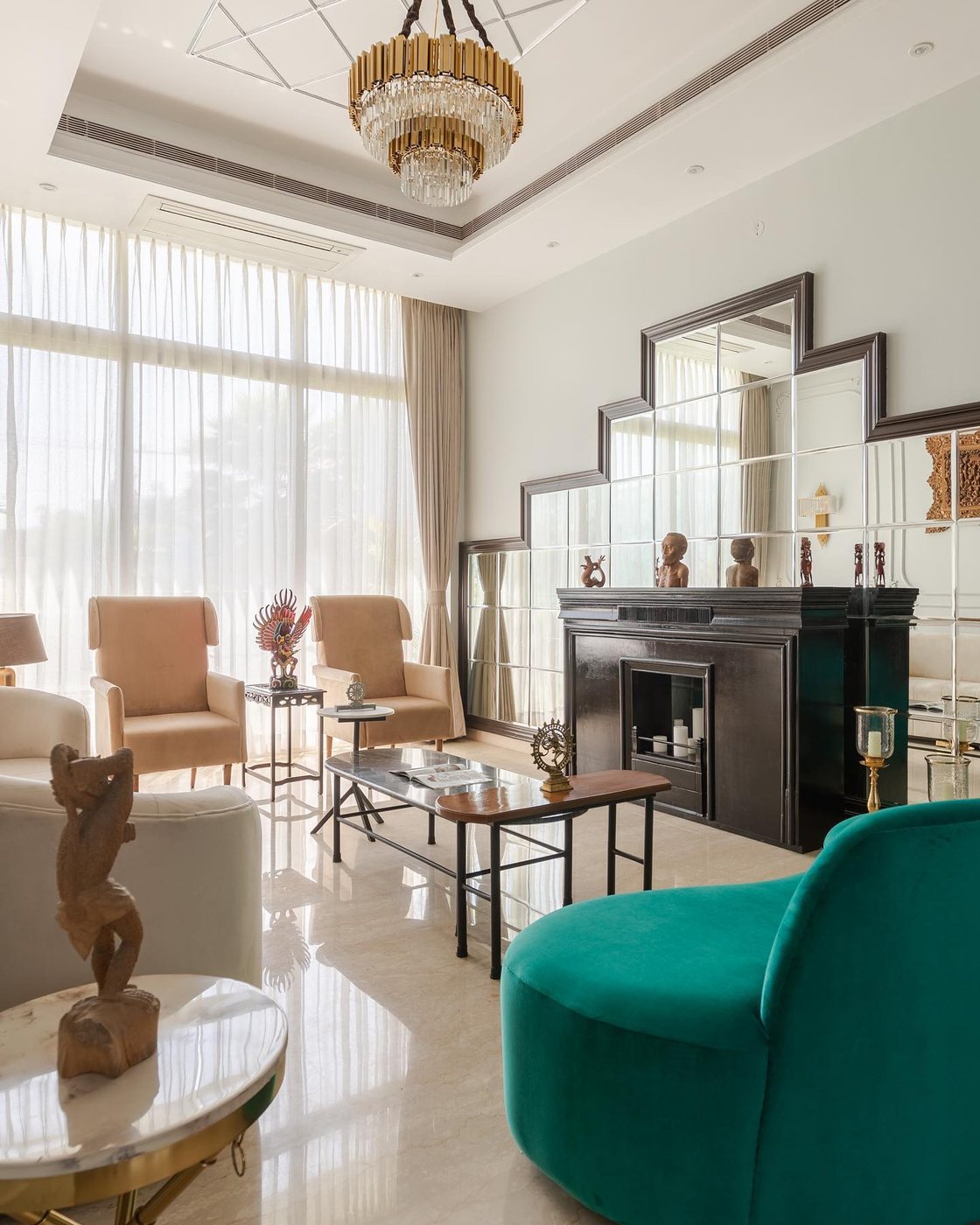 Living room with tiered mirrored fireplace surround, chandelier, and emerald velvet chair on marble floor
