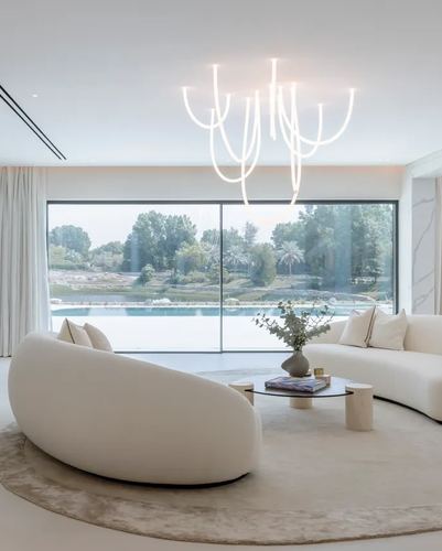 Curved cream sofa and round table under a sculptural chandelier facing a floor-to-ceiling window with pool view