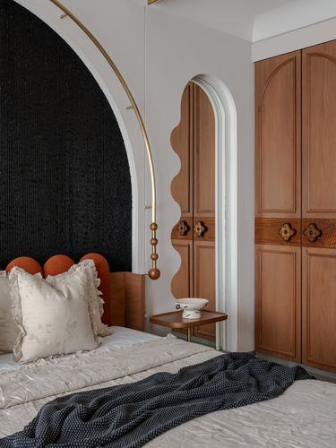 Bedroom with black arched textured headboard, terracotta bolsters, and carved wooden wardrobe beyond a scalloped archway