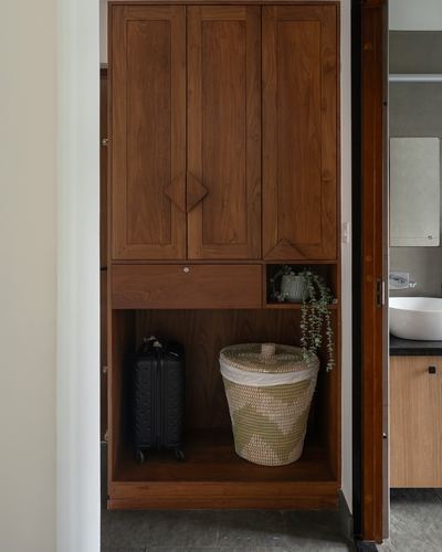 Wooden storage cabinet with open shelf holding a woven basket, plant, and suitcase, adjacent to bathroom door
