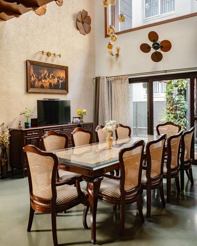 Dining room with ten cane-backed chairs around a long wood table under a double-height wall with decor.