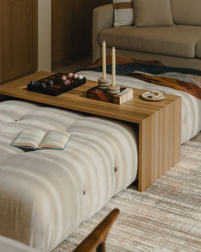 Wooden tray table spanning two linen tufted ottomans with candles and books, striped throw behind