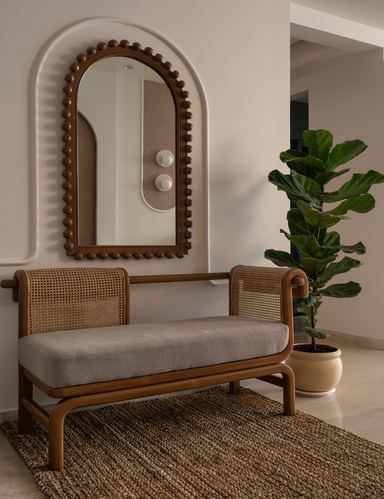Cane bench with grey cushion under arched mirror niche beside a leafy plant