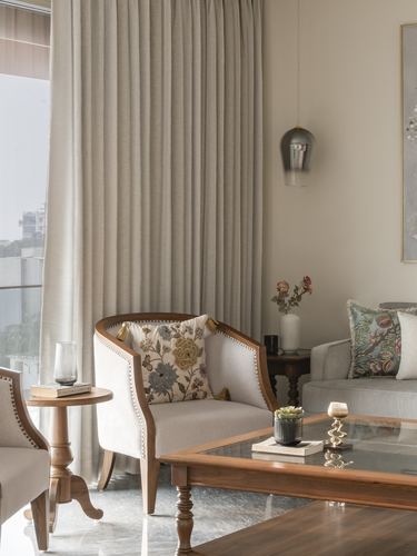 Living room corner with wood-framed armchair, floral cushion, glass pendant, and sheer curtains by window