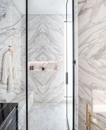 Book-matched marble shower with glass door, gold hook, and lit niche holding toiletries and a candle