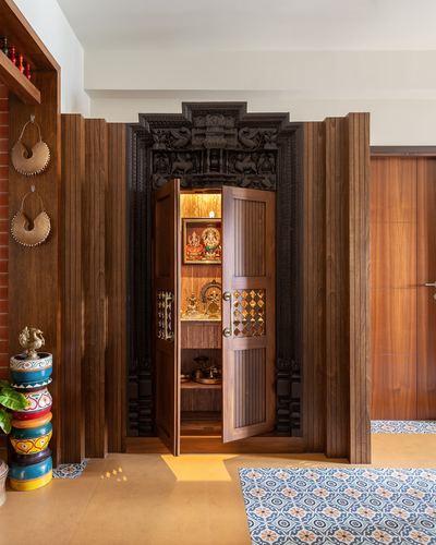 Open carved wooden pooja door revealing illuminated shrine with idols and brass lamps