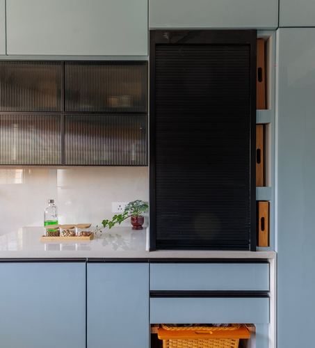 Pale blue kitchen cabinetry with ribbed glass upper cabinet and black tambour appliance garage, countertop styled with plant and jars.