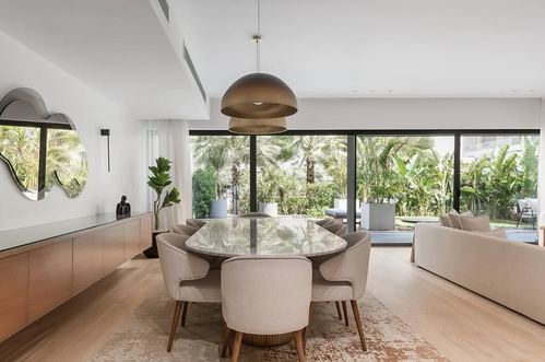 Neutral dining room with oval stone table, cane-backed chairs, and a bronze dome pendant facing floor-to-ceiling garden windows