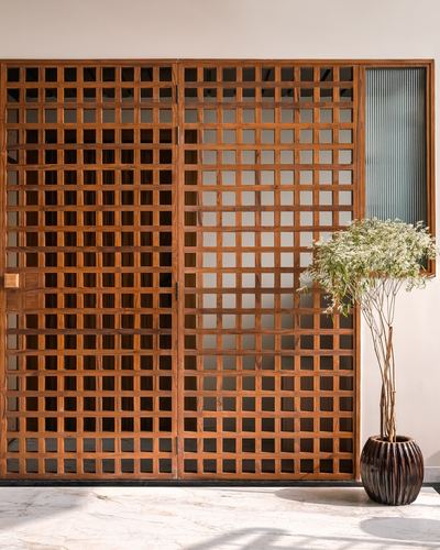 Wooden lattice entrance doors beside ribbed glass panel with tall potted plant on marble flooring
