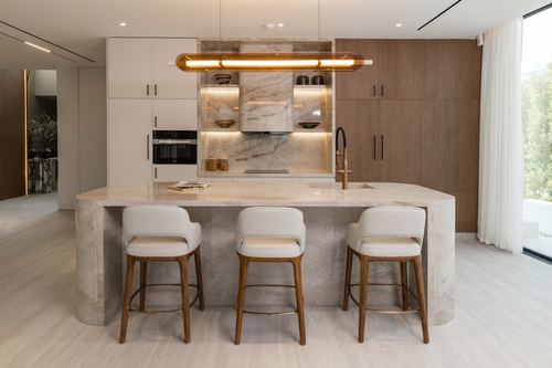 Contemporary kitchen island with white stools beneath a linear pendant light, stone backsplash niche behind cooktop