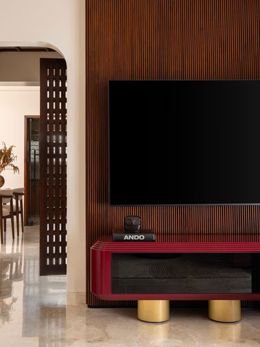 Living room with fluted wood wall, wall-mounted TV, and red lacquered console on brass legs beside a jali doorway