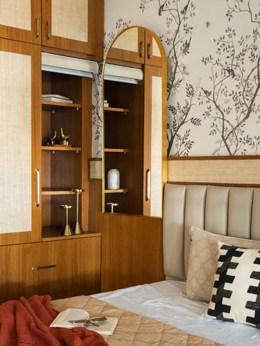 Wooden storage wall with arched mirror beside upholstered headboard and floral wallpaper