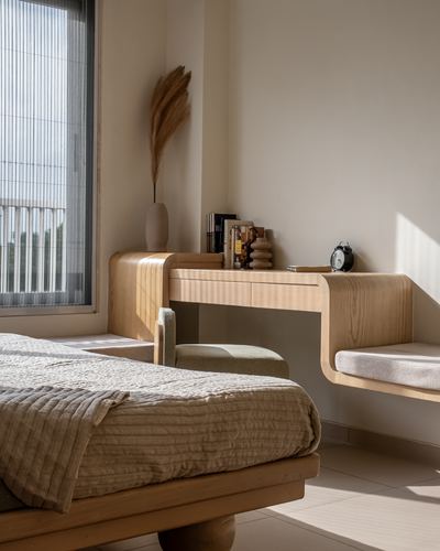 Sunlit bedroom corner with curved wooden desk, bench seat, and bed with striped linens.