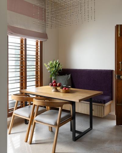 Dining nook with wooden table, purple velvet banquette, and geometric pendant light beside louvered windows