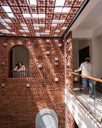 Woman leans from arched brick window as man walks past glass railing under a patterned terracotta skylight